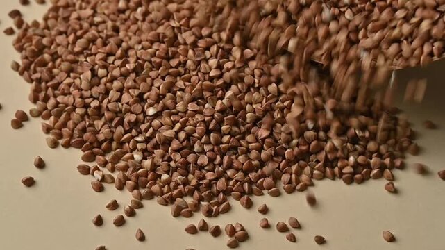 Close-up shot of dry organic buckwheat groats pouring from a wooden bowl onto a beige background. Healthy gluten-free food ingredient. 