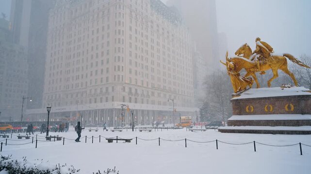Heavy snow is falling on the square, the William Tecumseh Sherman Monument and Plaza Park building. People walk by the street of New York City during blizzard.