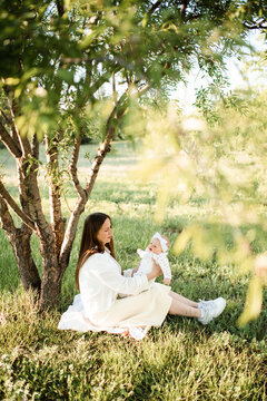 Happy woman holding baby sitting in green grass meadow under tree outdoors. Motherhood. Summer season.