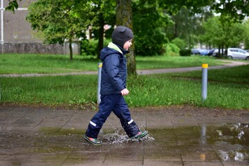 Fototapeta premium Toddler walking through rain puddles in waterproof outdoor clothing on a wet city sidewalk after spring rain. Playful childhood moment with splashing water and urban greenery in background.