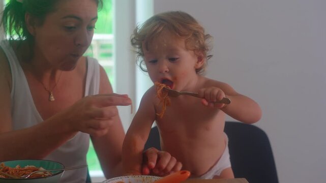 Mother helping toddler eat spaghetti during family mealtime while child feeds himself with fork capturing learning and authentic home parenting routine