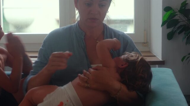Mother holding restless toddler while older child pleads for attention during busy family moment capturing multitasking and authentic everyday parenting life