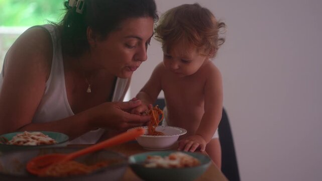 Mother feeding toddler spaghetti during family mealtime while child reacts with excitement capturing caregiving, nourishment and authentic home parenting moment