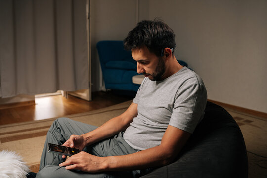 Man wearing bone conduction headphones relaxes at home, focused on mobile phone screen while streaming music and podcasts for private, comfortable listening experience.