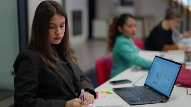 A young businesswoman writes notes and uses a laptop at her desk in a collaborative office. An employee works with focus and dedication during a busy workday.