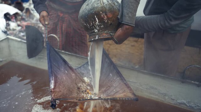 Natore, Bangladesh - 23 May 2026: View of workers pouring and filtering liquid through a cloth into a large metal vat during traditional gur production.