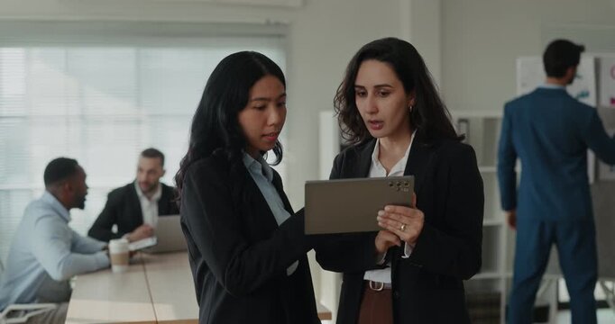 Portrait of Asian and Latin business women study tablet together as coworkers work near planning board. Colleagues review screen closely and discuss details