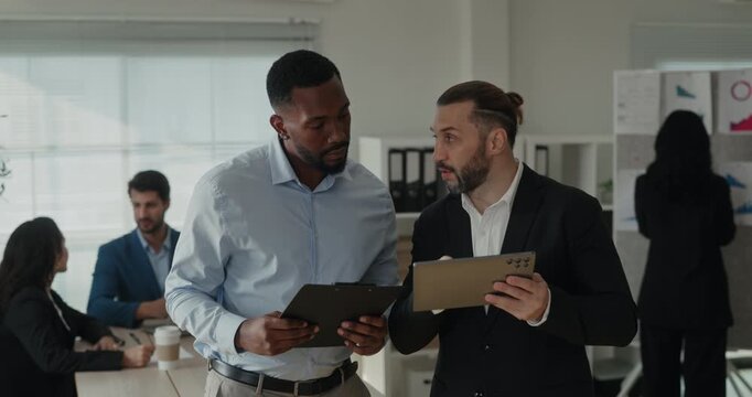 Portrait of Middle Eastern and African American businessmen review tablet data as team meets for briefing. Colleagues study screen closely and discuss report