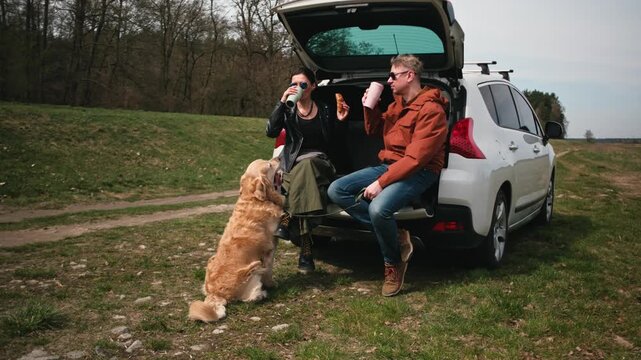 A couple relaxes at their car while eating snacks and drinking coffee. A golden retriever waits nearby on a grassy path in sunlight.