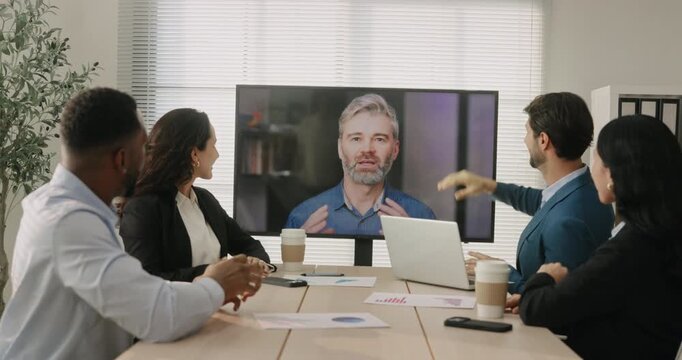 Multicultural business team greets remote manager on video call during online meeting in office. Coworkers wave at screen and welcome speaker on conference
