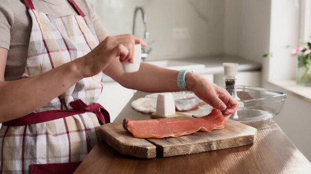 Woman in plaid apron seasons a salmon fillet on a wooden board. She sprinkles salt in a bright kitchen.