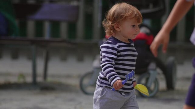 Toddler walking across playground sandbox carrying toy shovel and rake capturing independence, curiosity and authentic outdoor childhood exploration
