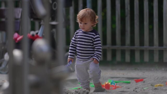 Toddler walking through playground sandbox while holding toy rake and balancing carefully capturing coordination, independence and authentic outdoor childhood play