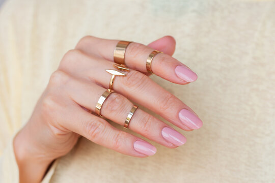 Woman's hand with a nude manicure, adorned with multiple gold rings on different phalanges of fingers, against a yellow background.