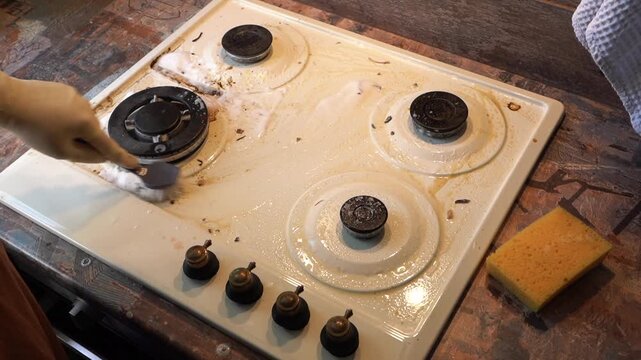 Gloved hand scrubbing greasy stovetop, closeup of white ceramic cooktop covered in burnt crumbs and oil, circular brushing motion with sponge and brush, soap foam pooling near burner, rustic