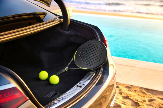 Black padel racket and two yellow balls in open luxury car trunk parked near beach pool. Padel sport equipment, summer lifestyle, active recreation concept.