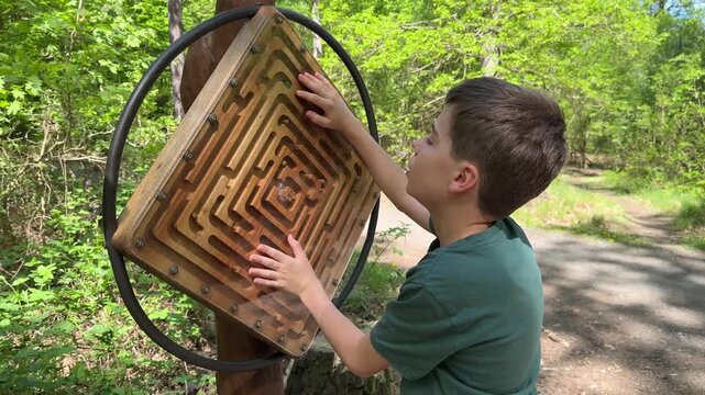 A child stands near a wooden maze in a park. He tries to solve the maze with his hands. The surrounding area has trees and a clear path. It is a bright day in spring.