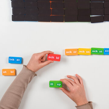 Top view of child's hands arranging colorful phonics word domino tiles on a white background. Educational literacy game for early reading skills.