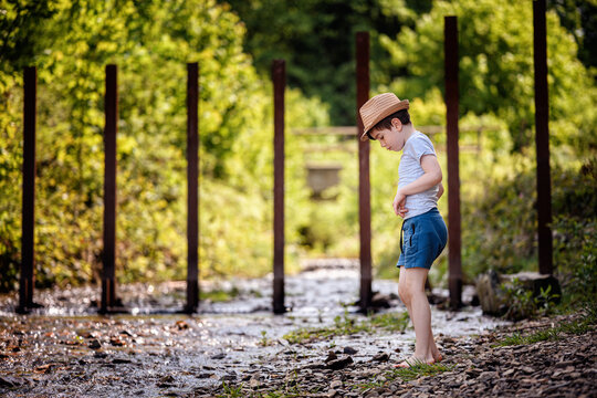 Thoughtful boy standing barefoot by shallow forest river looking down