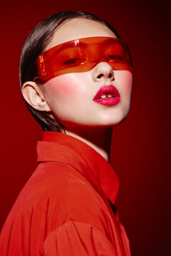 Stylish woman with bold red makeup and translucent orange visor strikes a fierce pose. Concept of beauty creative fashion with intense color. Studio portrait on red background.