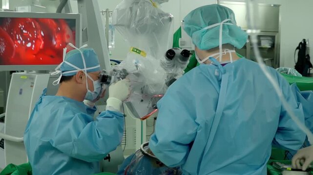 A medical professional performs a delicate procedure using an operating microscope in a surgical room surrounded by advanced monitoring equipment