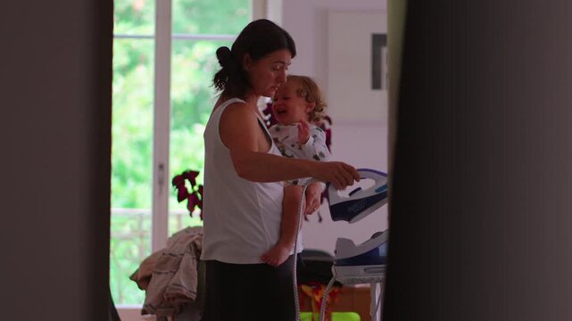 Mother holding toddler while ironing clothes interrupted by child seeking attention concept of multitasking parenting pressure and everyday family life