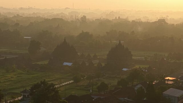 Aerial view of Plaosan Lor Temple shrouded in morning mist with golden sunrise light over the surrounding landscape in Kecamatan Prambanan, Jawa Tengah, Indonesia.