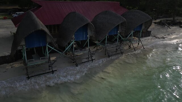 Aerial view of beach huts with thatched roofs on a sandy shore by the turquoise sea in Riau Islands, Indonesia.