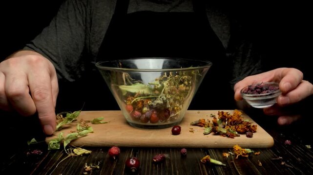 Hands preparing herbal mixture in glass bowl, adding dried flowers and berries on wooden cutting board with scattered ingredients around