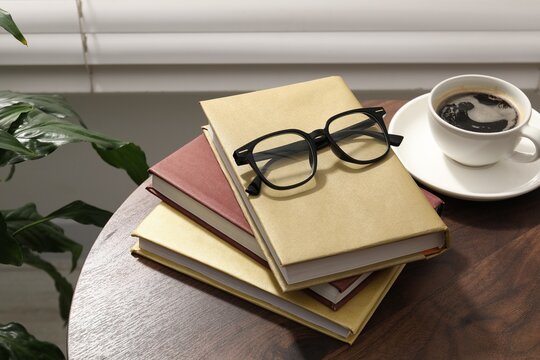 Pair of stylish glasses, books and cup of coffee on wooden table indoors, closeup