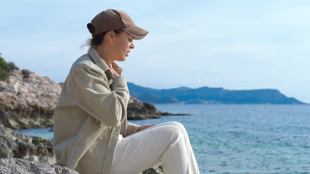 Woman using asthma inhaler near the sea. Tourist woman managing sudden asthma attack while sitting on rocky coastline, using inhaler during seaside vacation with blue sky background