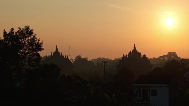 Aerial view of Plaosan Lor Temple silhouetted against a golden sunset sky with surrounding trees and rural houses in Kecamatan Prambanan, Jawa Tengah, Indonesia.