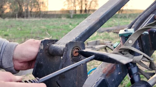 Close-up of male hands using a metal hammer to remove a steel pin from tractor loader machinery during farm equipment repair. Authentic agricultural maintenance scene with manual labor, mechanical
