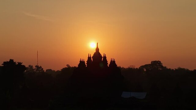 Aerial view of the silhouette of Plaosan Lor Temple against a golden sunset sky with trees in Kecamatan Prambanan, Jawa Tengah, Indonesia.