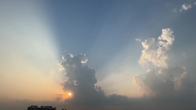 A dramatic evening sky featuring powerful crepuscular rays bursting upward from behind a dark cloud formation during sunset, with a faint silhouette of building rooftops visible along the horizon.