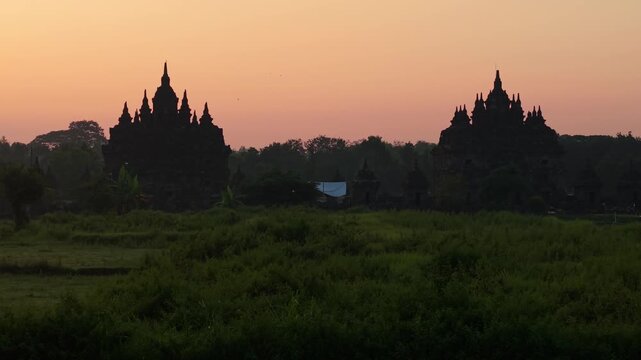 Aerial view of the silhouetted Plaosan Lor Temple twin structures against a vibrant sunset sky over lush green fields in Kecamatan Prambanan, Jawa Tengah, Indonesia.