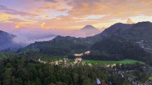 Aerial view of Dieng Plateau featuring terraced fields and misty mountains under a colorful sunset sky in Kecamatan Batur, Jawa Tengah, Indonesia.