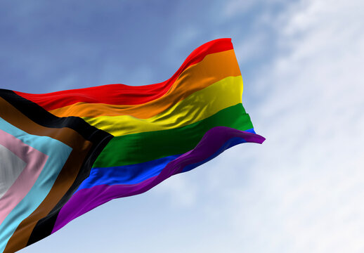 Progress Pride Flag ripples in the wind against a blue sky with soft clouds. 3D rendered illustration of the inclusive LGBTQ+ symbol for diversity and equality.