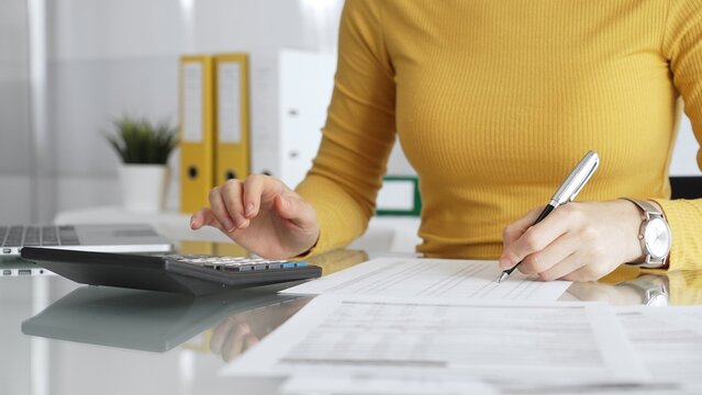 Businesswoman wearing yellow casual sweater is calculating budget and reviewing financial documents using a laptop and calculator for home finances. Audit and taxes in business