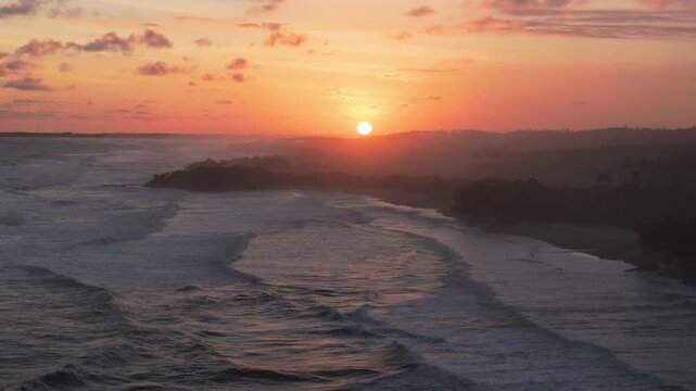 Aerial view of the sunset over Ujung Genteng Beach with waves crashing on the shore under a vibrant orange sky in Kecamatan Ciracap, Jawa Barat, Indonesia.