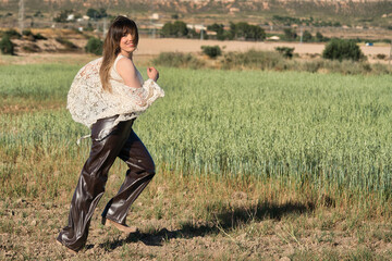 Fototapeta premium Woman leaps joyfully through green. Her laughter echoes as she runs barefoot. Sunlight bathes the field in golden warmth. She wears a lace top and glossy pants. 