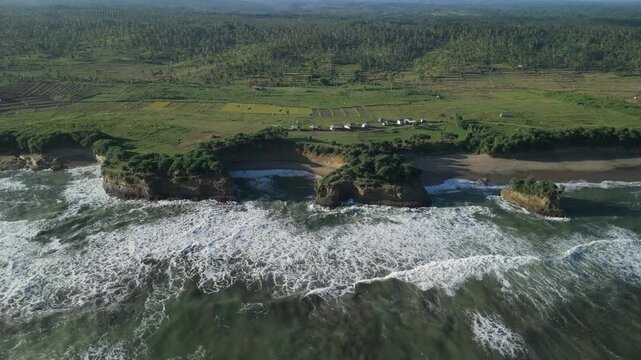 Aerial view of Ujung Genteng Beach with crashing waves against rocky cliffs and lush green fields in Kecamatan Ciracap, Jawa Barat, Indonesia.
