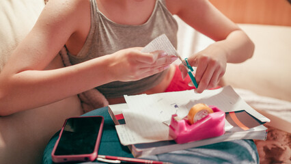 Close up of a focused student hand using adhesive tape and scissors on a small piece of paper, finishing a secret crib sheet and notes for a test while sitting on a sofa in a living room. © Hektor2