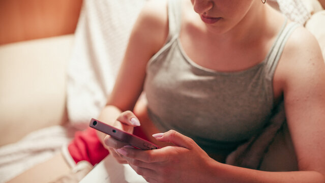 Focused high school girl sitting on a couch and typing on her mobile phone, using a smartphone to copy notes, browse exam answers, or cheat on a future test while studying at home.