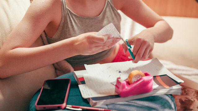 Close up of a focused student hand using adhesive tape and scissors on a small piece of paper, finishing a secret crib sheet and notes for a test while sitting on a sofa in a living room.