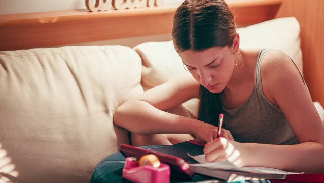 Deeply focused and worried high school girl making secret notes and tiny cheat sheets from her mobile phone, preparing for a test while sitting on a couch under warm indoor lighting.