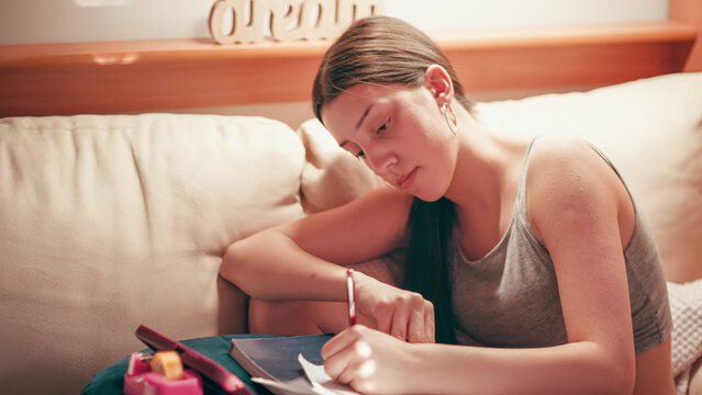 Deeply focused and worried high school girl making secret notes and tiny cheat sheets from her mobile phone, preparing for a test while sitting on a couch under warm indoor lighting.