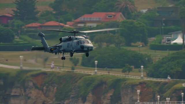 Gray naval military helicopter with landing gear deployed flies at low altitude over a coastal cliff and park, sensor equipment visible on fuselage