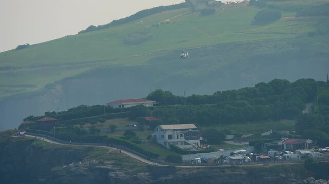 White military helicopter flies in hazy distance above a scenic coastal headland, winding road and cluster of houses with campervans visible below