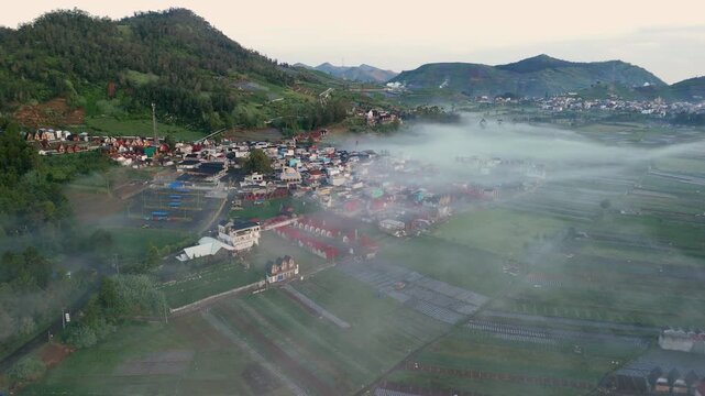 Aerial view of Dieng Plateau with morning mist rolling over a village and terraced fields in Kecamatan Batur, Jawa Tengah, Indonesia.